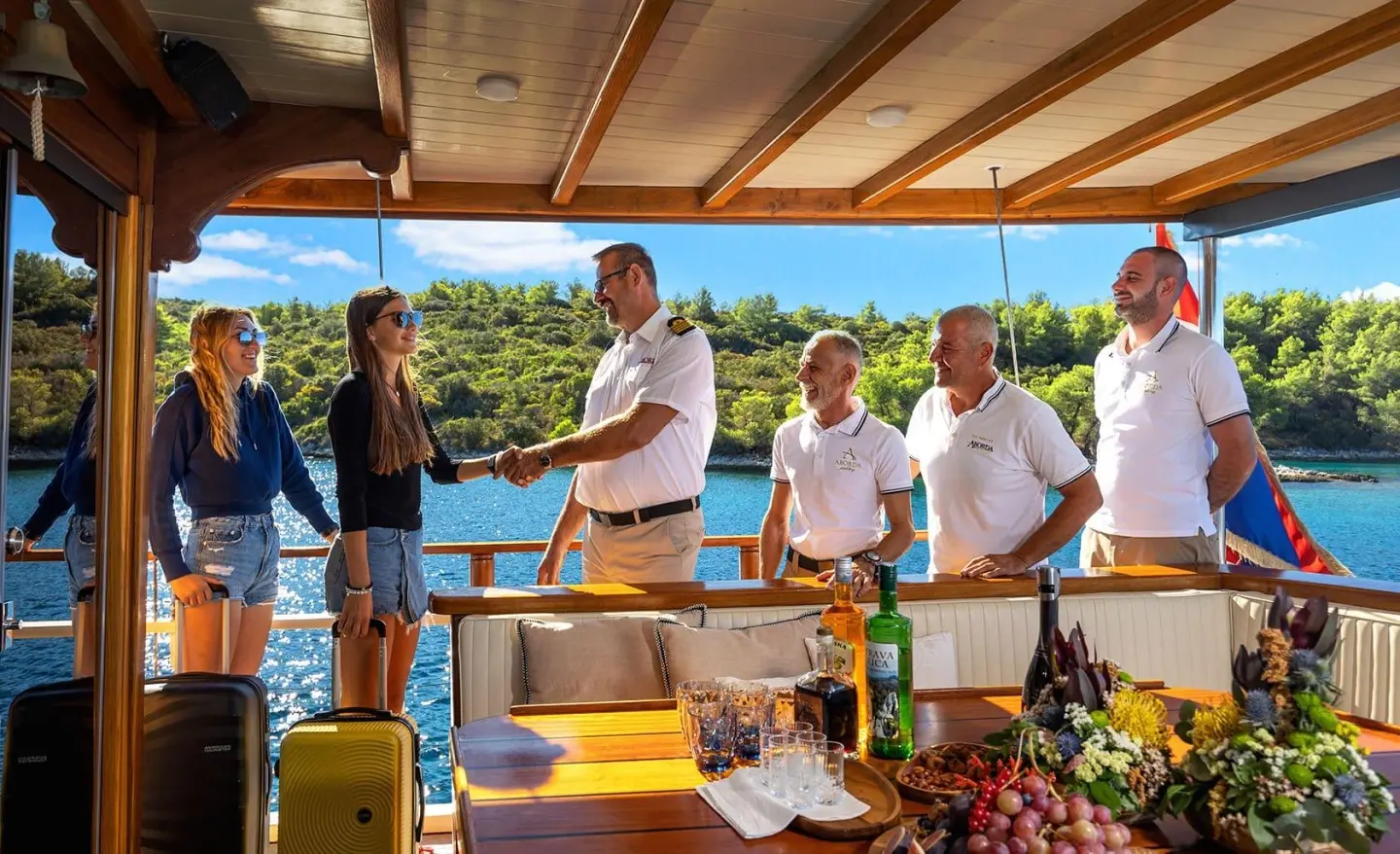 Yacht Aborda crew on deck with passengers under a wooden cabin, sunny marina scene