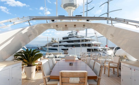 Long wooden table with seating on a yacht deck, loungers and boats in the background under blue sky