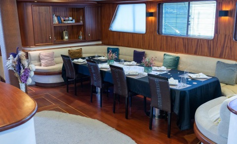 Nevra Queen yacht interior dining area with long table, chairs, wood paneling, and large windows with blinds