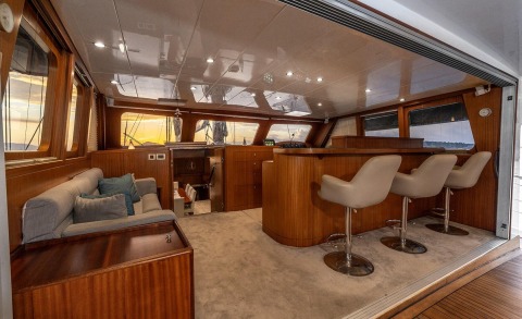 Wood-finished interior of a yacht with a bar and beige chairs, sunset light through windows