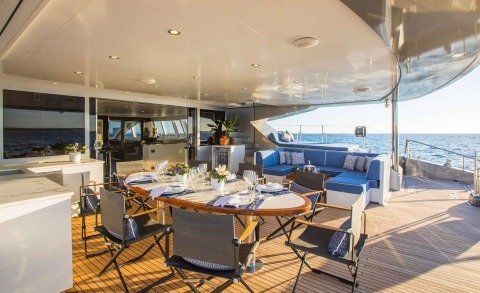 Open deck with a luxurious motor yacht dining area around a wooden table, chairs and stools; blue and white sofa seating and the sea visible in the background