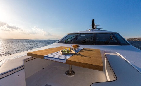 On the yacht deck a wooden table with a striped cloth, fruit and drinks, sea and sunlit sky in the background