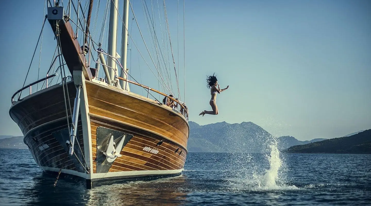 Entre Cielos yacht with wooden hull sailing near a rocky cliff at sunset