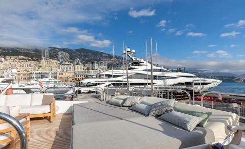 Large luxury yacht deck with cushioned lounge area; masts and a docked yacht in the background