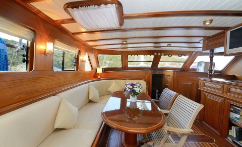 Wood-paneled interior of a yacht with cream seating, a polished wooden table, and side windows showing the outside scenery