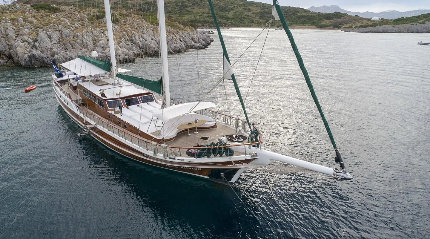 Ecce Navigo luxury schooner at anchor near rocky coastline, teak deck and green sail cover