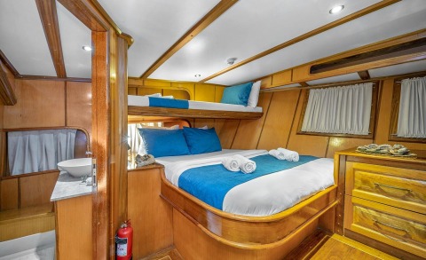 Wooden yacht interior with lower and upper beds, blue pillows, white sheets, and wooden cabinets with curtains