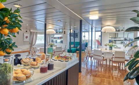 La Perla luxury yacht interior: bright salon with dining area, fruit display, pastries, and teal seating details