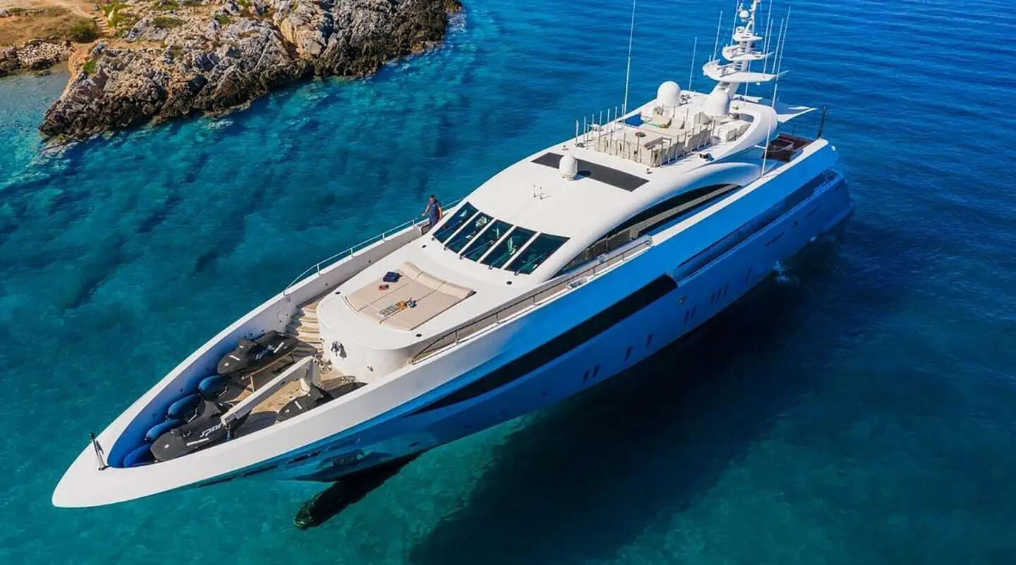 Large white luxury motor yacht anchored in clear blue water near rocky shore