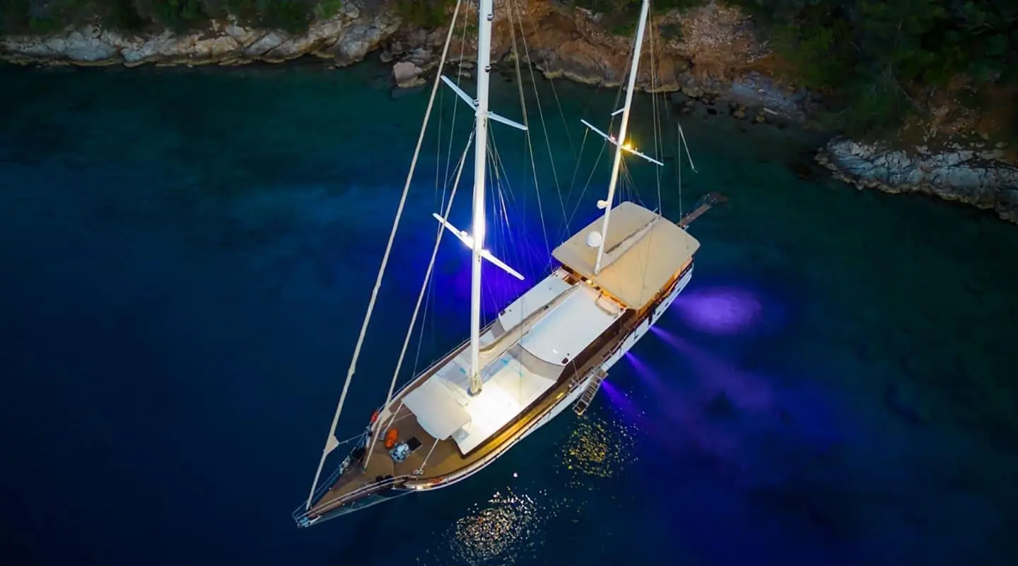 Gulet yacht in night lighting on dark blue water beside rocky shore