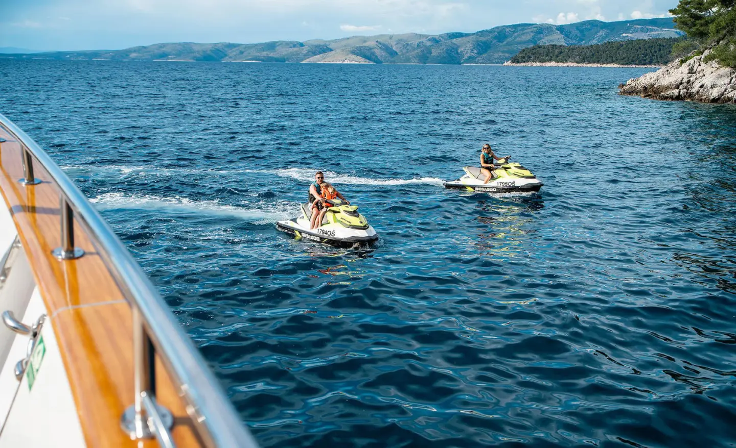 Two people riding yellow-green jet skis on blue open sea near rocky coastline