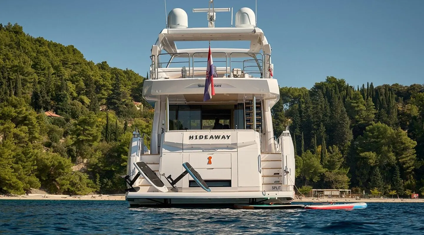 Back view of a multi-deck luxury yacht named Hideaway docked by trees on calm water.