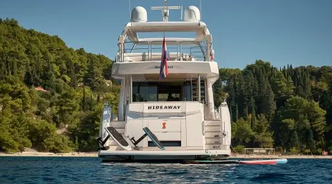 Back view of a multi-deck luxury yacht named Hideaway docked by trees on calm water.