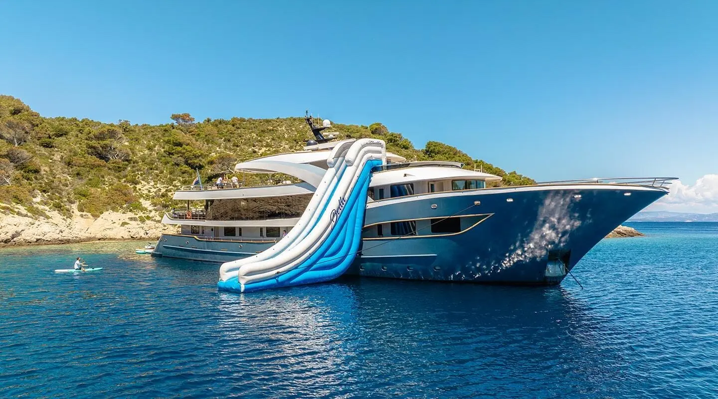 Bella yacht with blue waterslide at stern, docked near hillside coastline