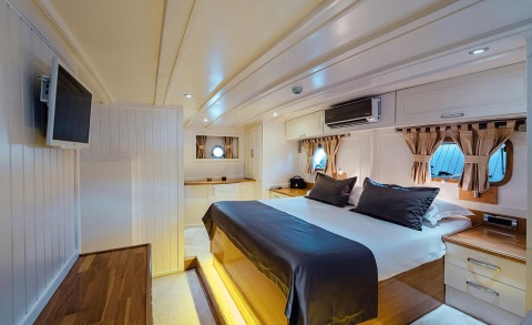 Interior of a yacht cabin with white wood paneling, large bed with navy and white bedding, and windows with curtains