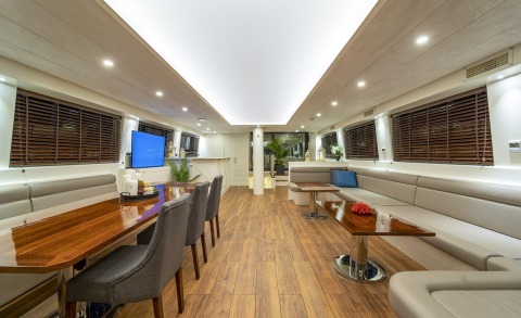 Interior salon with long wooden dining table and white seating, boat interior with blinds on windows