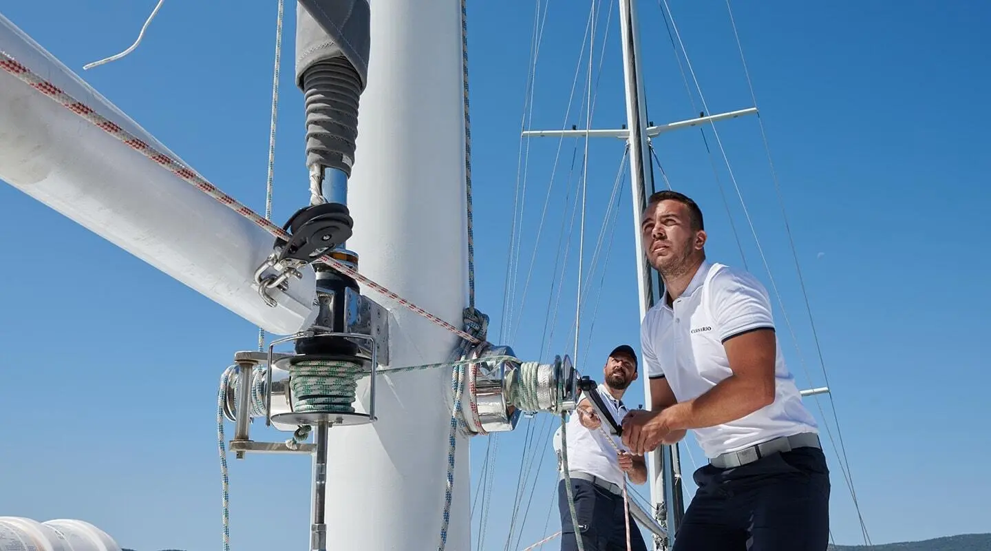 Two crew members working on a sailboat mast and rigging under bright blue sky