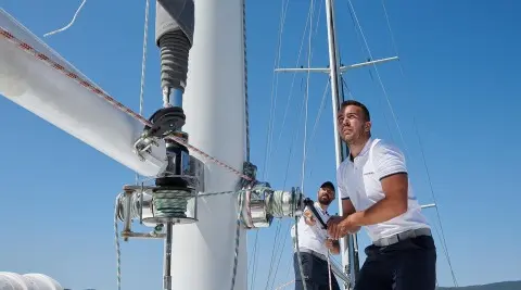 Two crew members working on a sailboat mast and rigging under bright blue sky