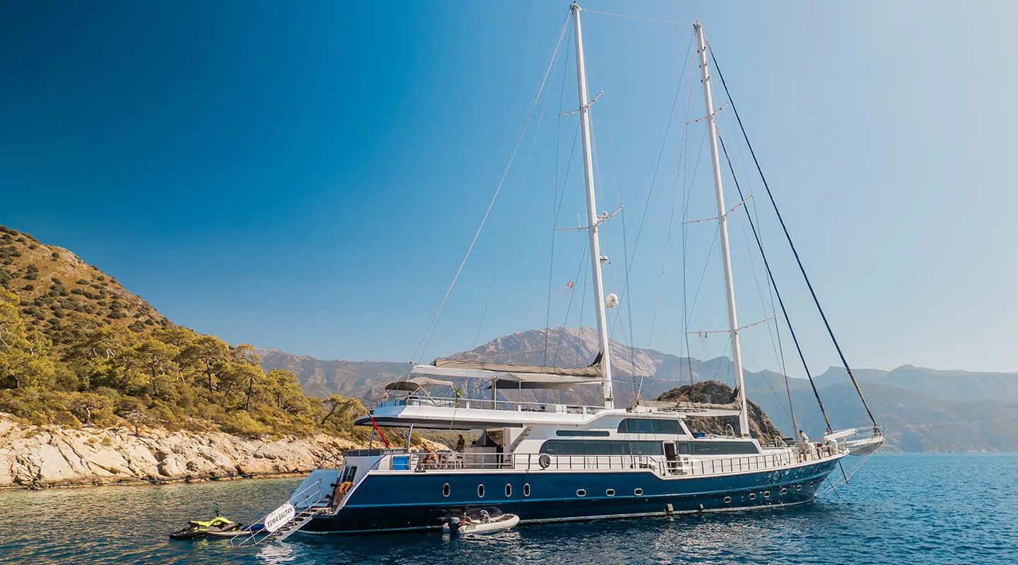 Gulet Esma Sultan anchored near rocky coast with masts and blue sky