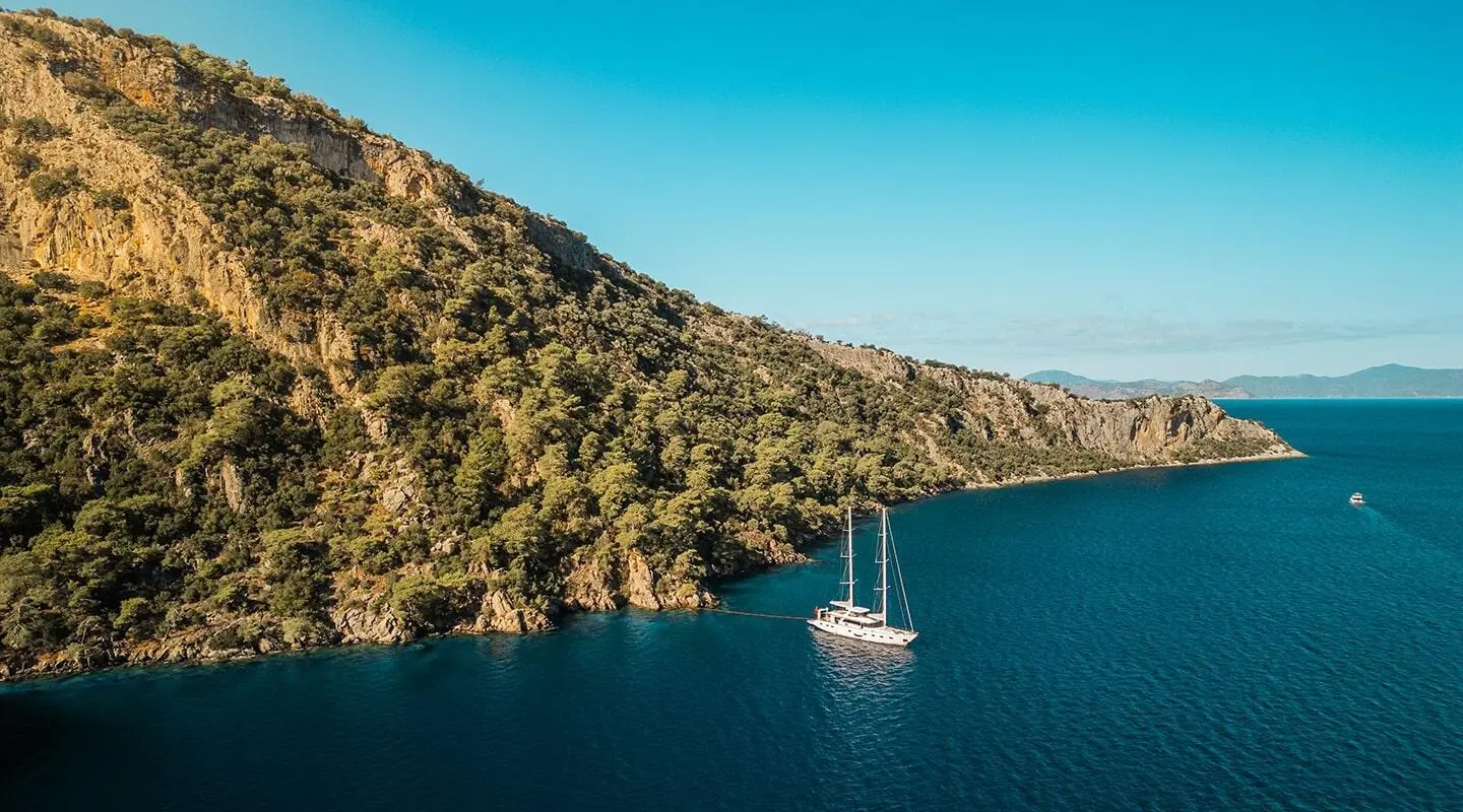 A schooner-style luxury yacht anchored in a calm cove near rocky coastline with blue sea