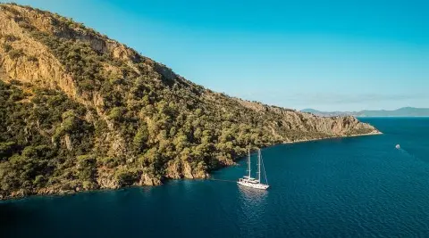 A schooner-style luxury yacht anchored in a calm cove near rocky coastline with blue sea