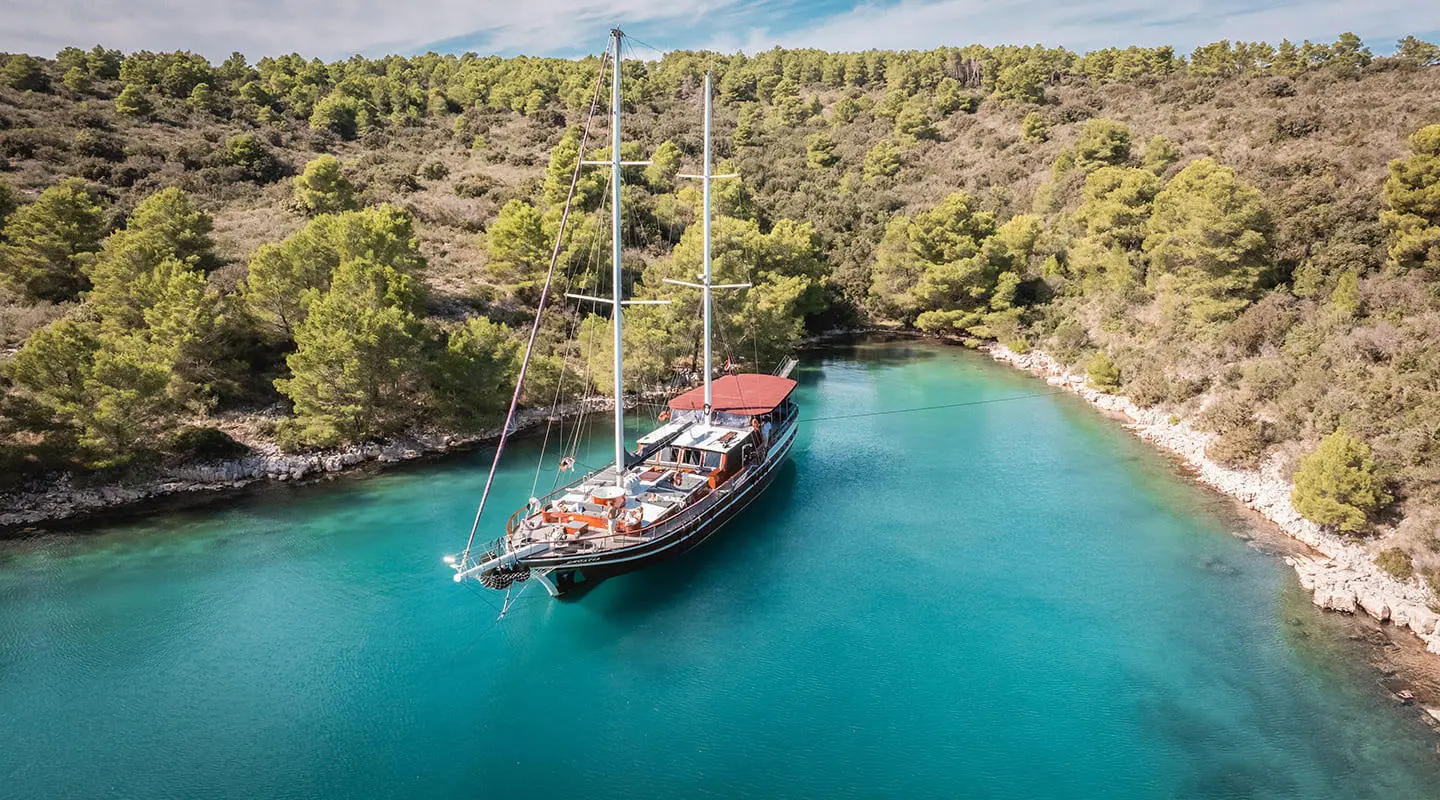 Double-mmast gulet anchored in a turquoise cove by a forested shore