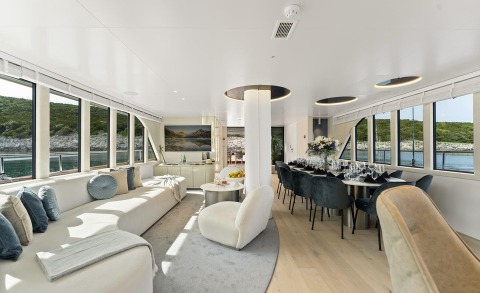 Maxita yacht interior: bright lounge with cream sofas, round dining table, large windows and coastal view