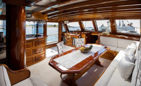 Andi Star’s polished wooden interior with a central dining table and helm, overlooking blue sea through large windows