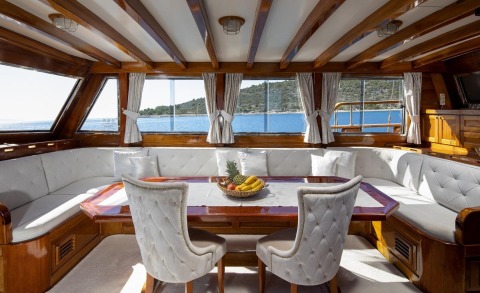 Wooden interior salon with white upholstered seating and a table, windows showing sea and shore outside