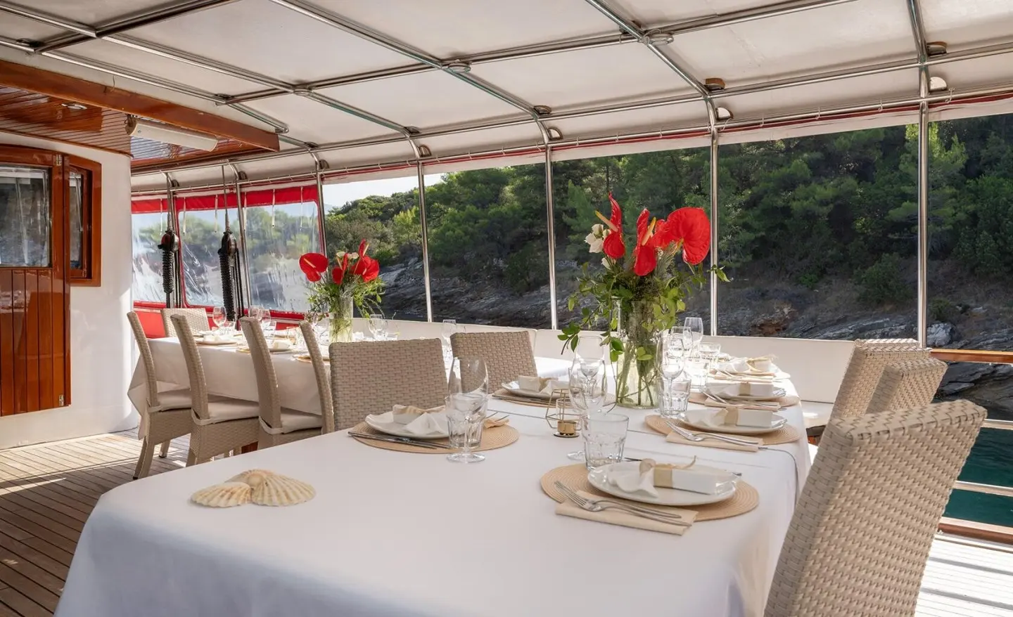 Table set for a meal on a yacht under a canopy, with red flowers and china