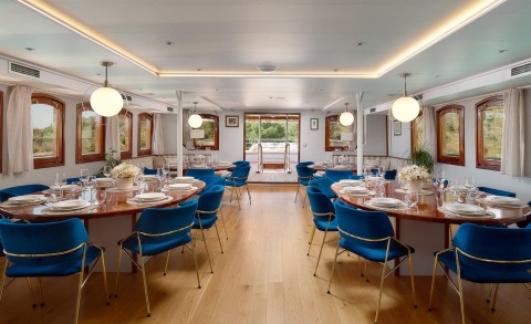 Dining area aboard with blue chairs around wooden tables, windows along sides, and ceiling lights