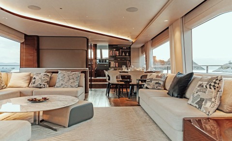 Vista motor yacht salon with long cushioned sofas, a dining table, bookshelves, and large windows showing the sea