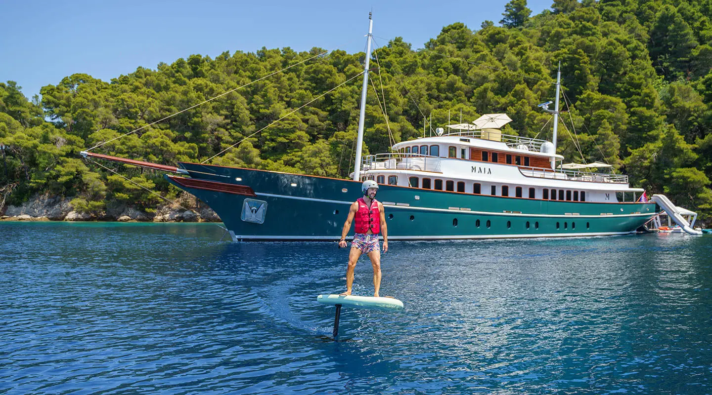 Maia yacht anchored near a rocky coastline with a paddleboarder in foreground