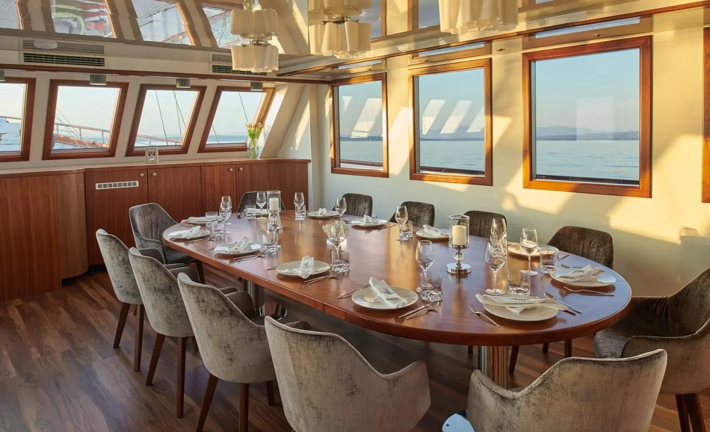 Sunlit wood table and chairs inside a yacht dining area with windows overlooking the sea