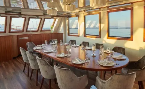 Sunlit wood table and chairs inside a yacht dining area with windows overlooking the sea