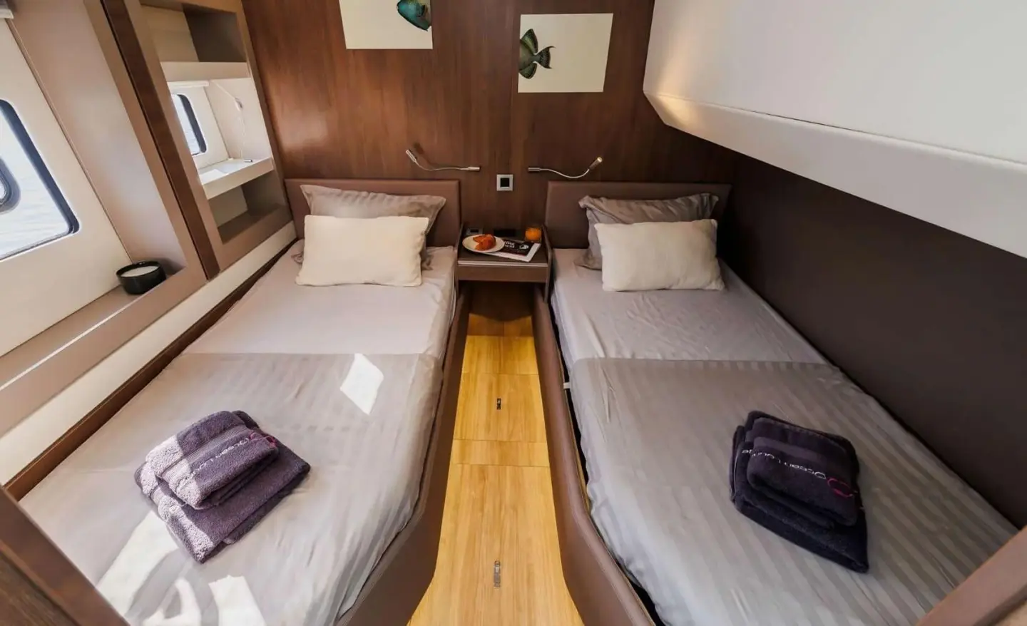 A teak cabin interior with two single beds, a central wooden aisle, and a small cabinet on the left.