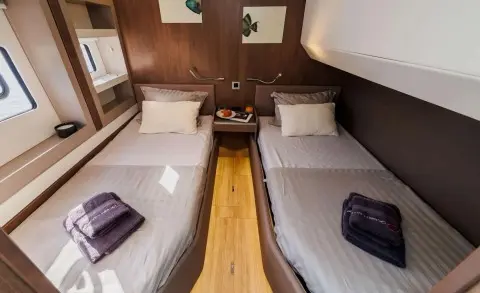 A teak cabin interior with two single beds, a central wooden aisle, and a small cabinet on the left.