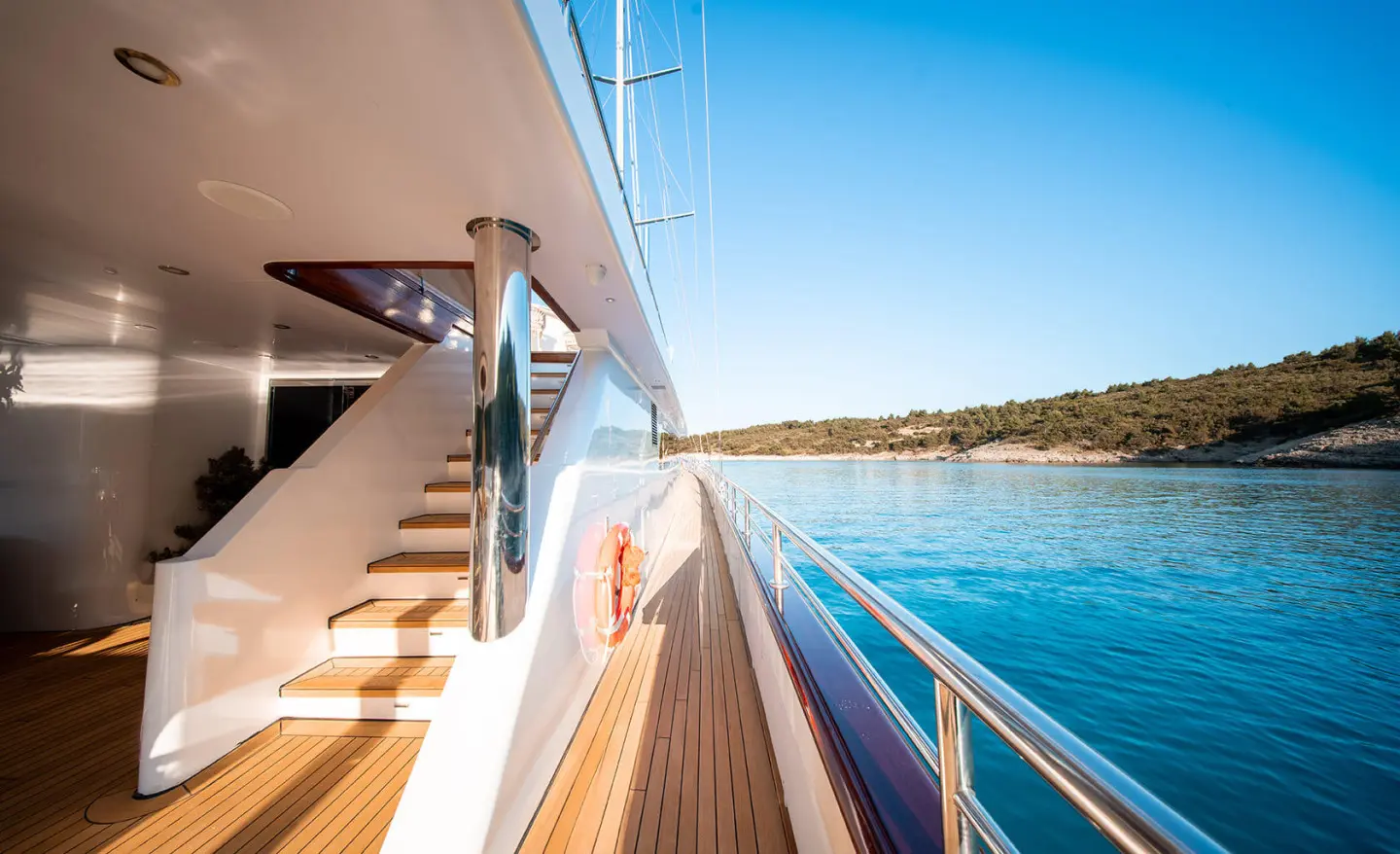 Side deck of Nocturno motor yacht, sunlit polished wood and railings by clear blue water