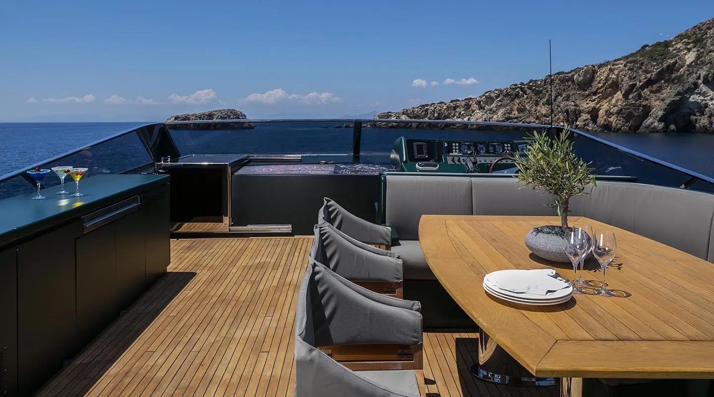 A luxury yacht deck dining area with a long wooden table and black chairs overlooking blue sea and coastline