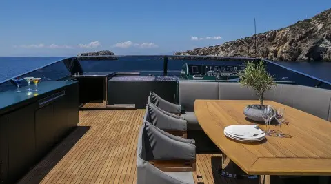 A luxury yacht deck dining area with a long wooden table and black chairs overlooking blue sea and coastline