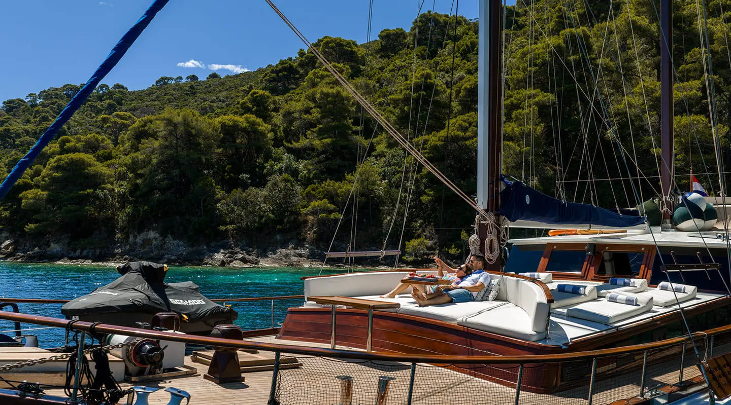 Elegant yacht with white hull and sails docked at a sunny marina, pine-covered hills in background