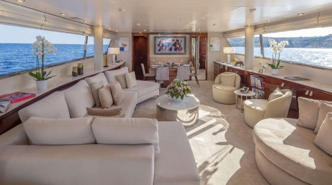 Interior of a yacht lounge with white sofas and a round coffee table; dining area in the back and sea view through windows