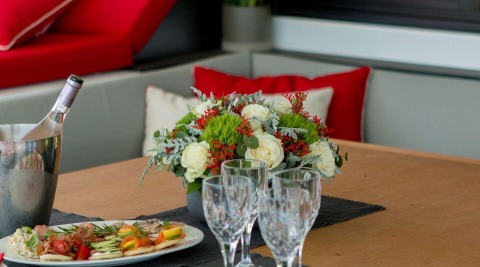 Wooden table with floral centerpiece and champagne bucket, plates of appetizers and crystal glasses visible inside a yacht lounge