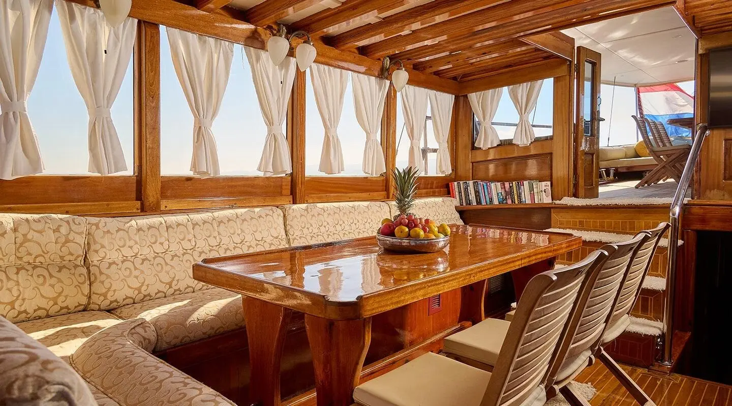Linda yacht charter interior: wooden seating, tables, beige cushions, sunlit windows, sunset view