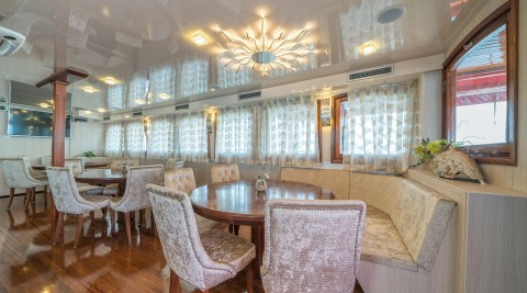 Round wooden table with cream upholstered chairs in the Barbara yacht salon, white patterned curtains on the windows