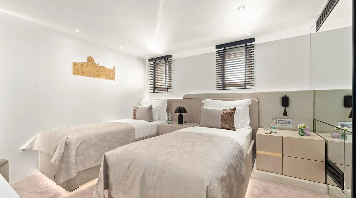 Two side-by-side twin beds in a bright yacht cabin with beige bedding and white walls