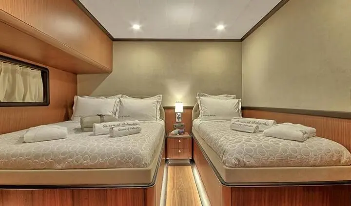 Queen Of Salmakis yacht cabin with twin beds, warm wood accents, bedside lamps