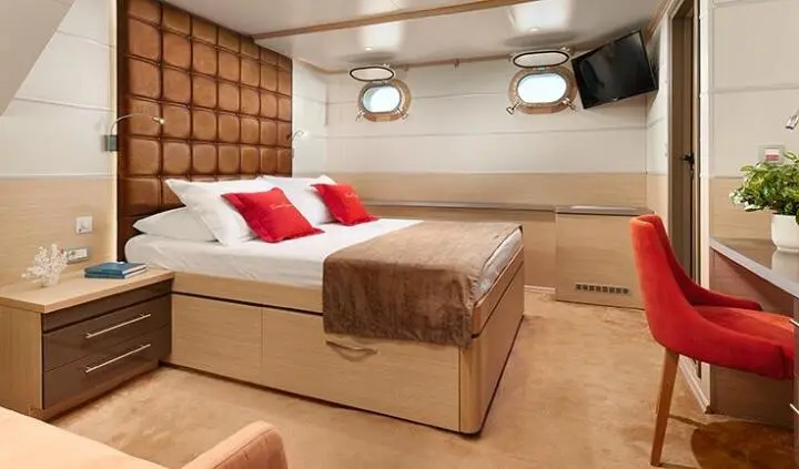 Queen Eleganza cabin bed with brown blanket, red pillows, and porthole window view