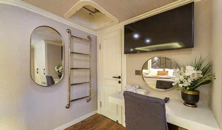 Queen Of Makri cabin interior with wall mirror, mounted TV, desk, and plush chair