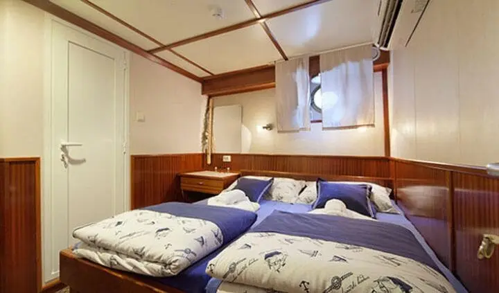 Interior cabin with two single beds joined, wooden walls, blue bedding, small window, overhead cabinet